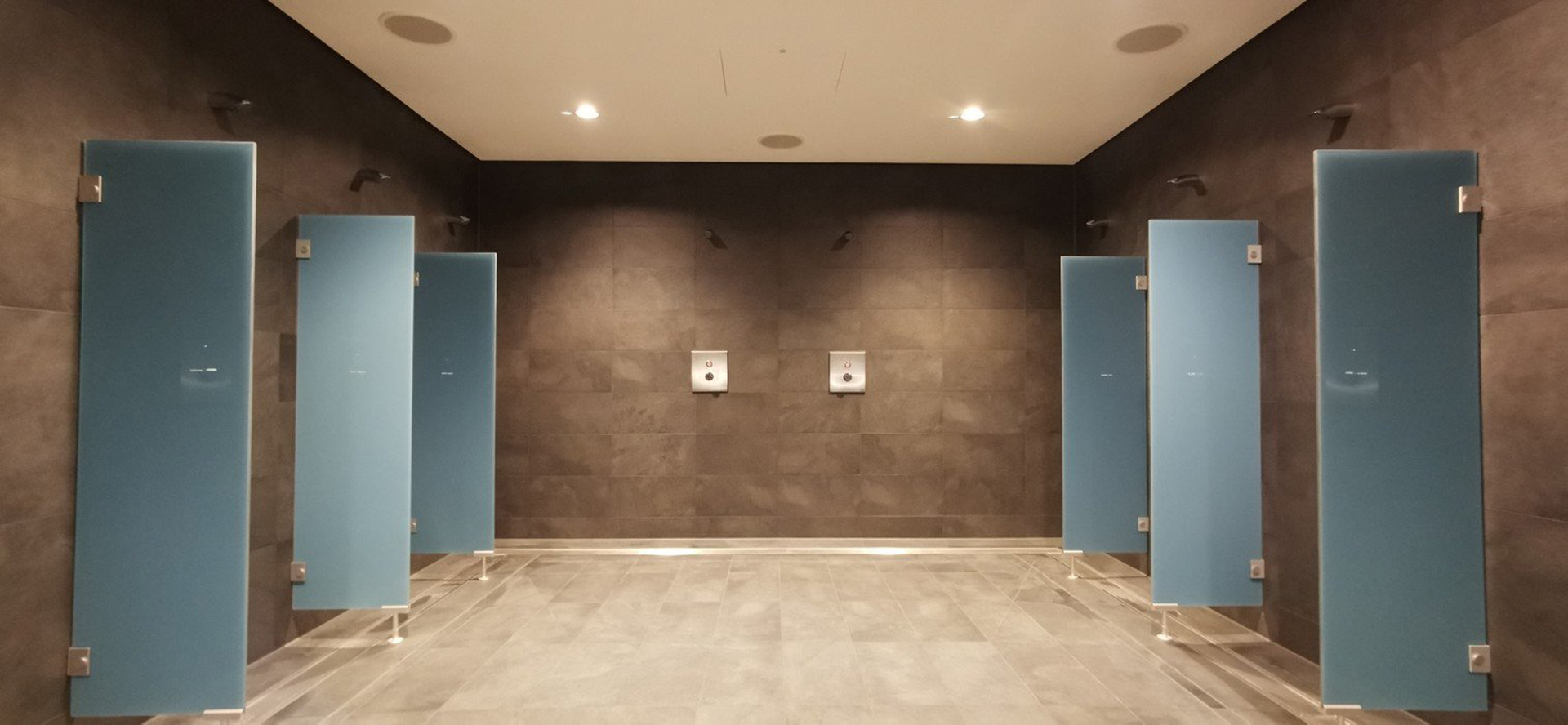 Moderner Duschbereich im Hallenbad mit grauen Fliesen, breitem Durchgang und blauen Glastrennwänden.