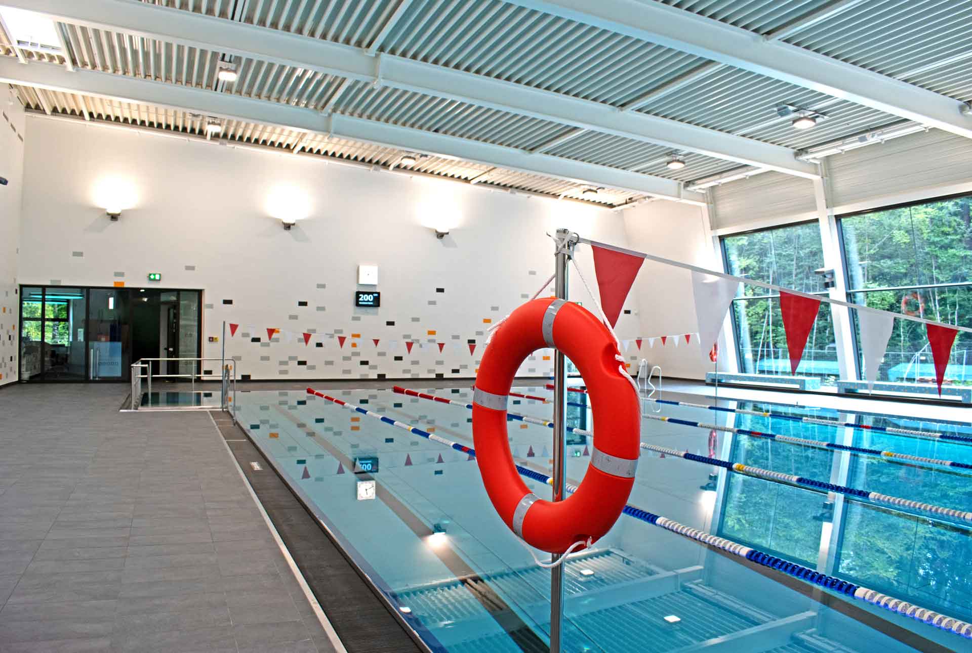 Roter Rettungsring vor den Schwimmbahnen eines modernen Hallenbads mit großen Fenstern und Startblöcken.
