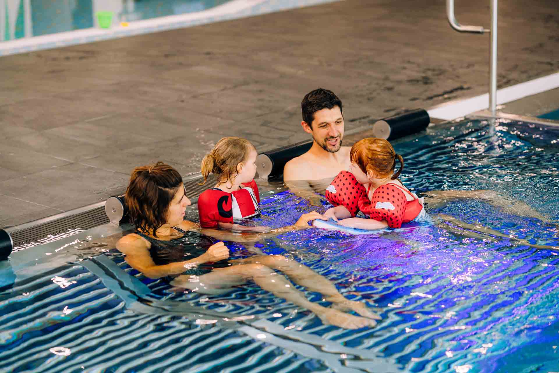 Zwei Erwachsene begleiten zwei Kinder mit roten Schwimmhilfen im flachen Becken eines Hallenbads.