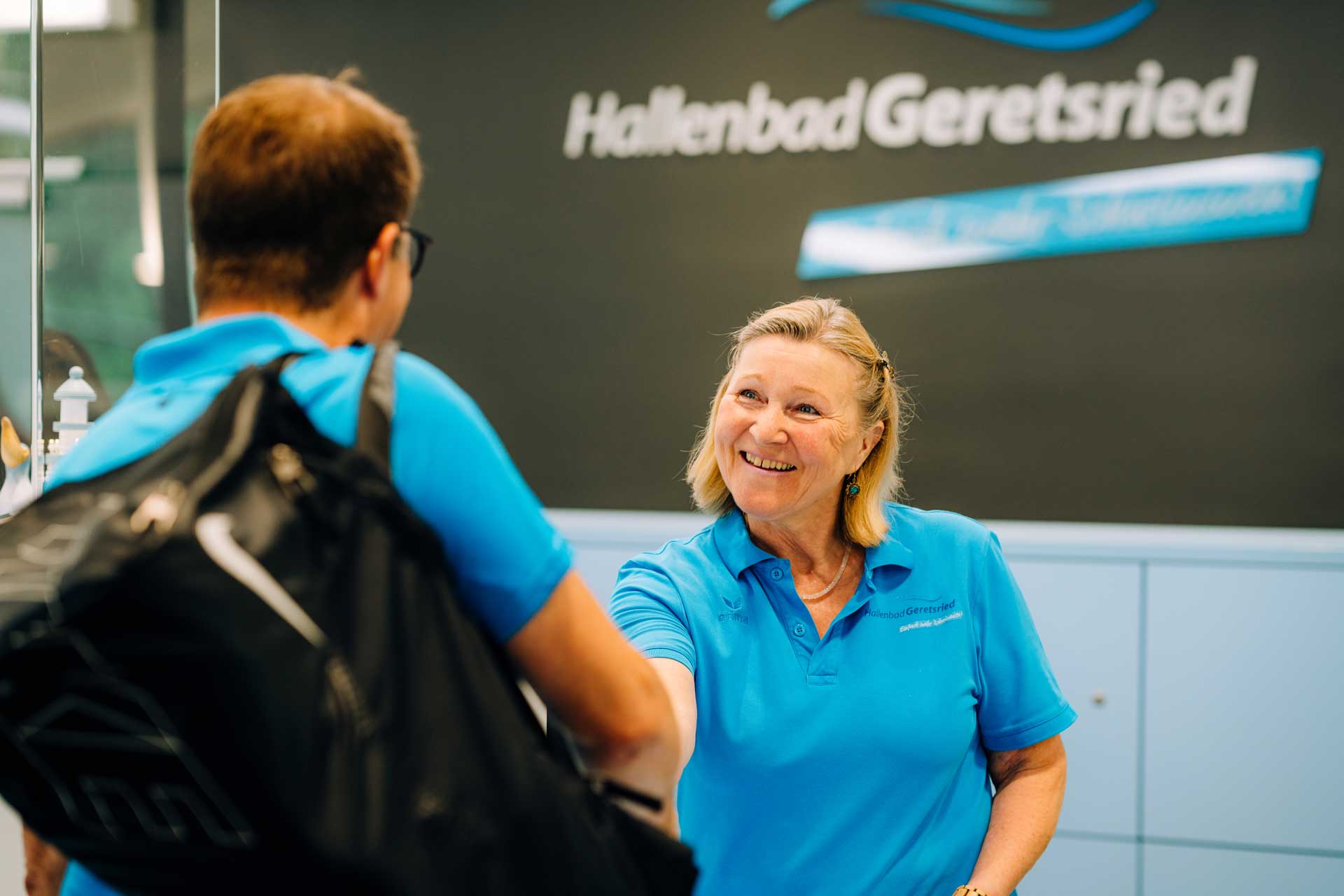 Lächelnde Mitarbeiterin des Hallenbads Geretsried begrüßt einen Besucher mit Rucksack am Empfang.