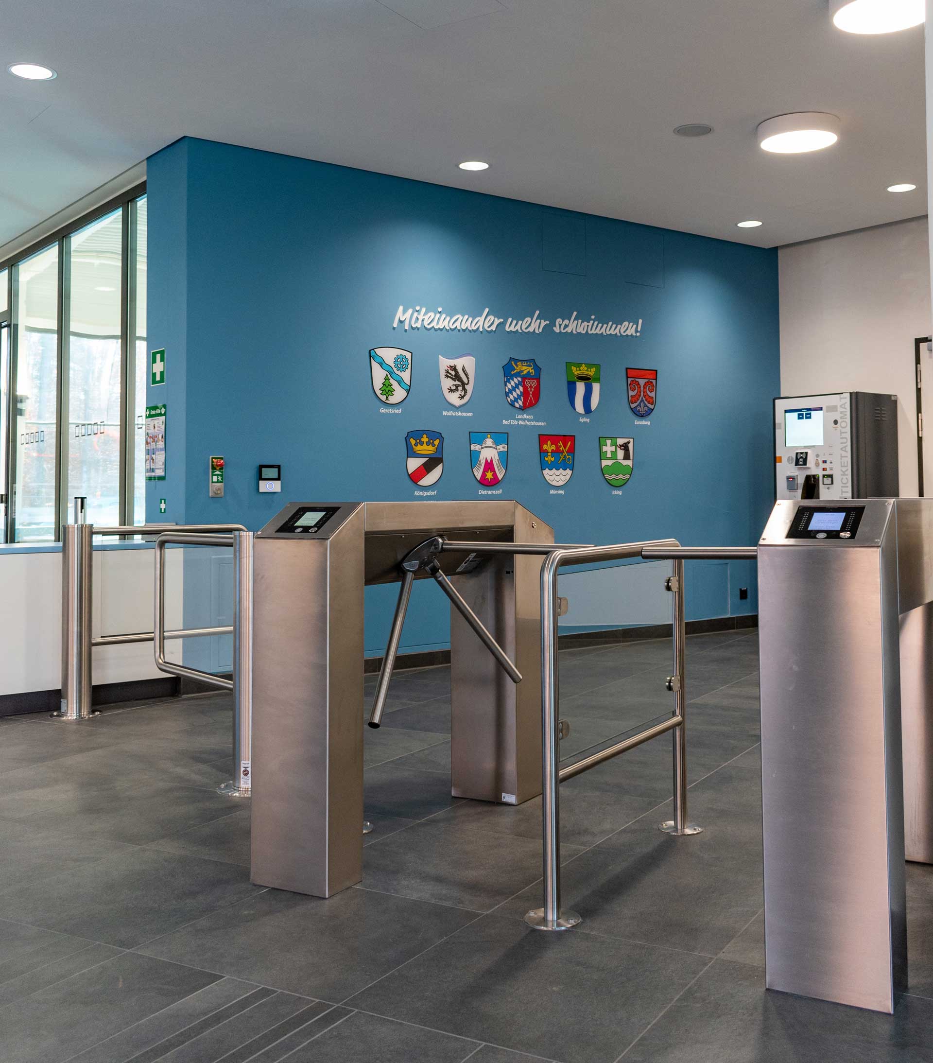 Eingangsbereich des interkommunalen Hallenbads mit Drehkreuzen, Ticketautomat und Wappen der beteiligten Kommunen.