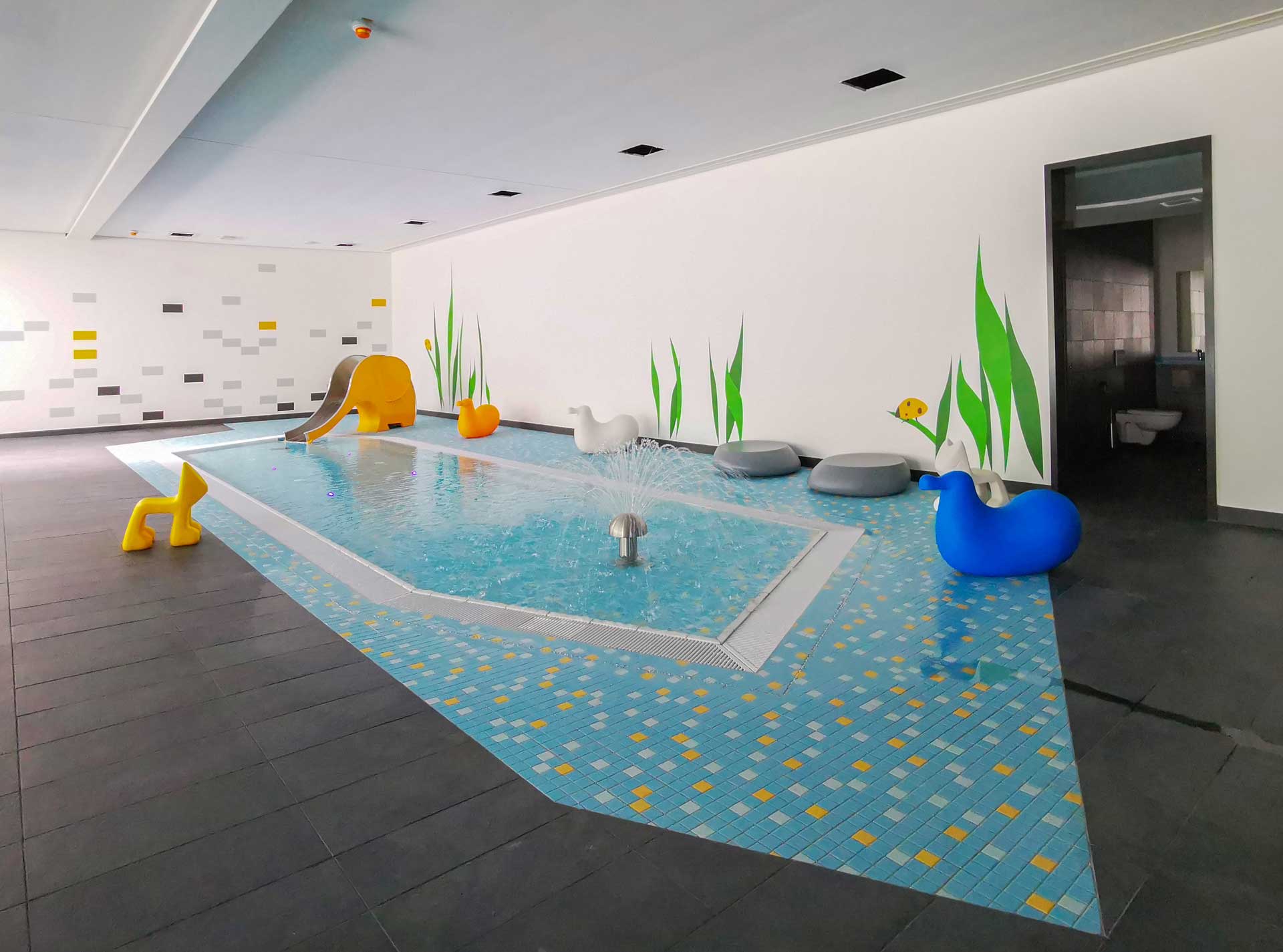 Kinderplanschbecken mit Wasserfontänen, gelber Elefantenrutsche und bunten Tierfiguren im Hallenbad.