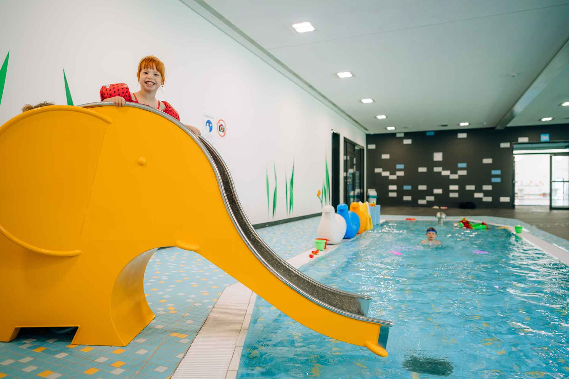 Lachendes Kind rutscht auf einer gelben Elefantenrutsche ins Kinderplanschbecken des Hallenbads.