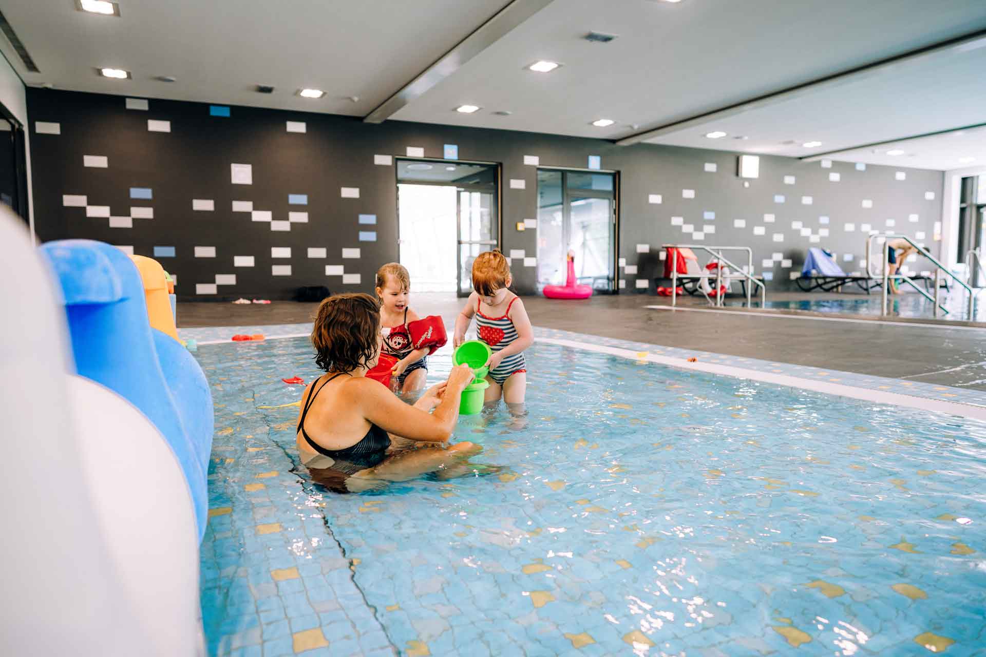 Zwei Kinder spielen mit einem grünen Eimer im flachen Kinderplanschbecken, begleitet von ihrer Mutter.