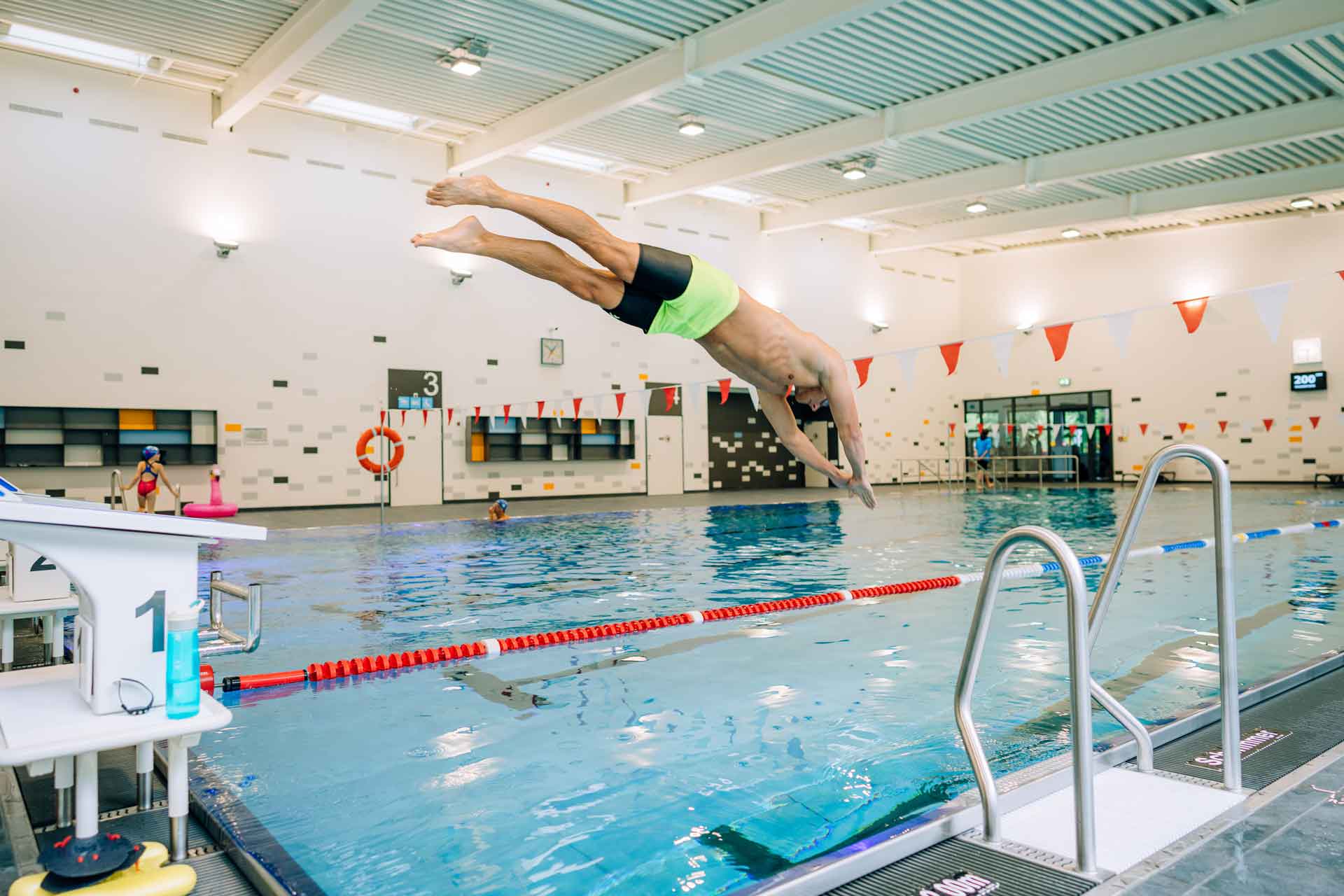 Schwimmer springt kopfüber vom Startblock in ein abgetrenntes Becken eines hellen Hallenbads.
