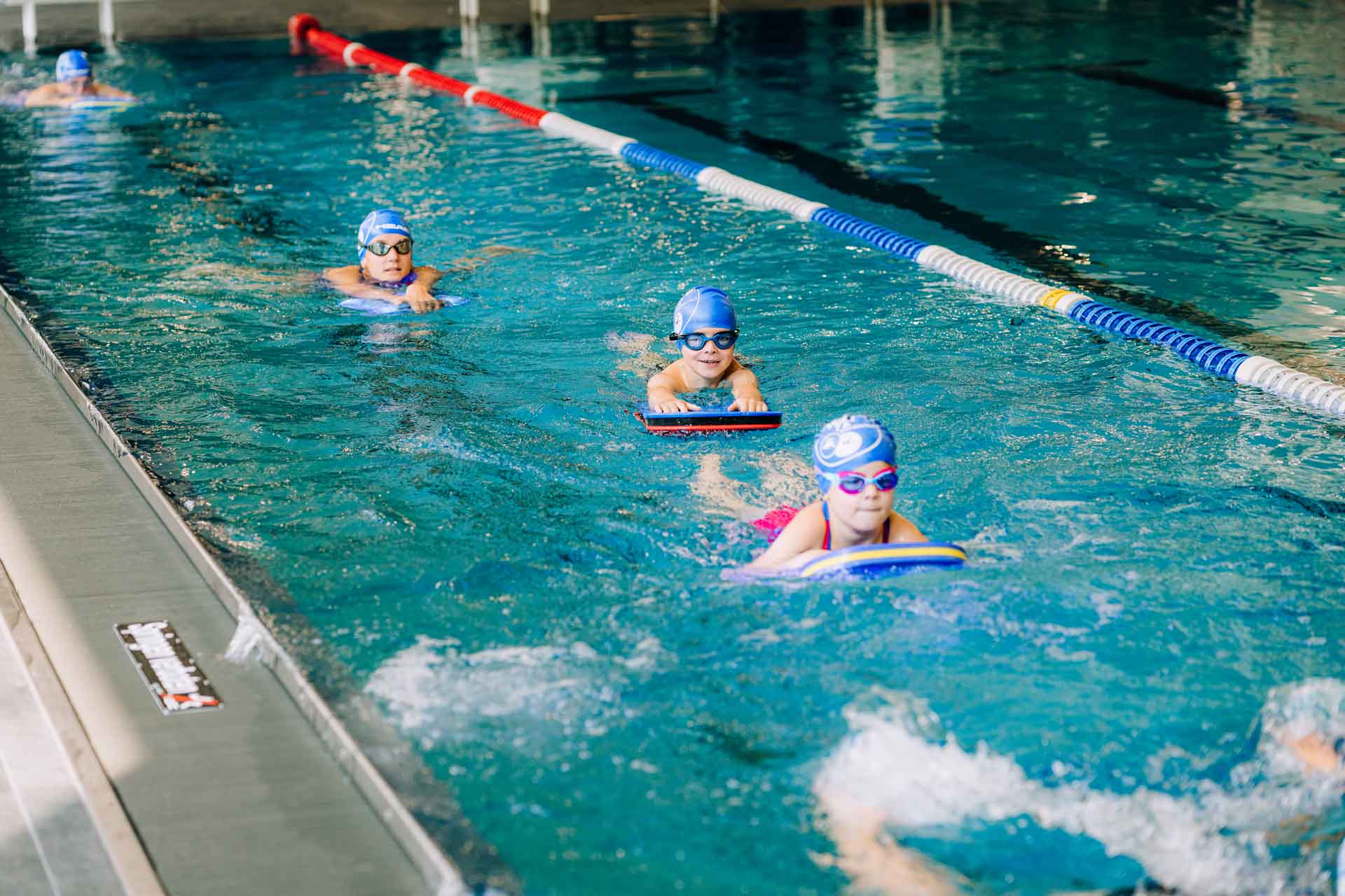Kinder mit Badekappen und Schwimmbrillen üben mit Schwimmbrettern in einer Bahn im Hallenbad.