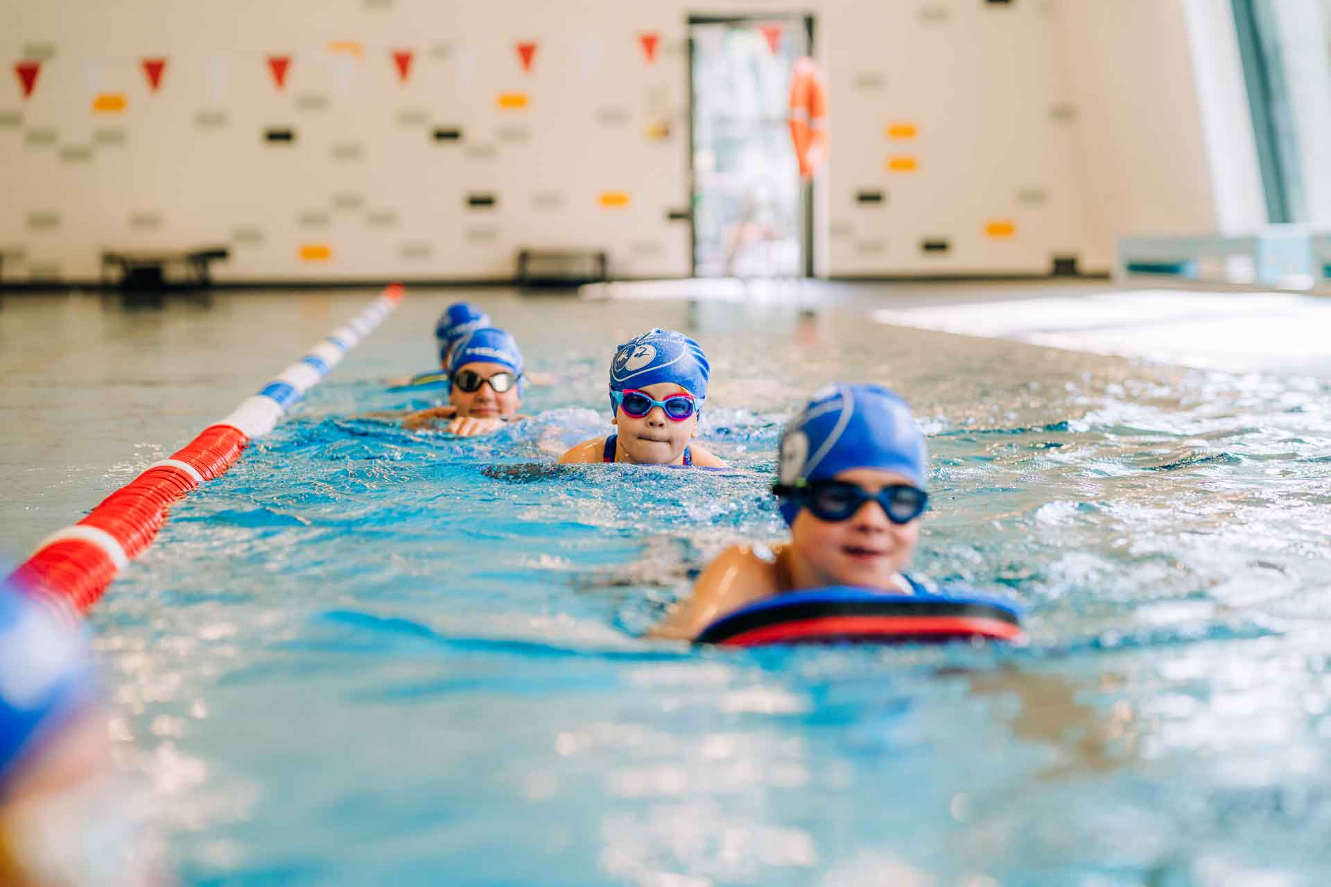 Drei Kinder mit blauen Badekappen und Schwimmbrillen trainieren hintereinander im Hallenbad.