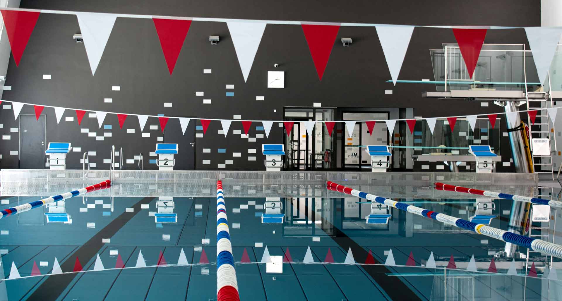 Schwimmerbecken im Hallenbad mit fünf Startblöcken, roten, weißen und blauen Leinen sowie Sprungturm.