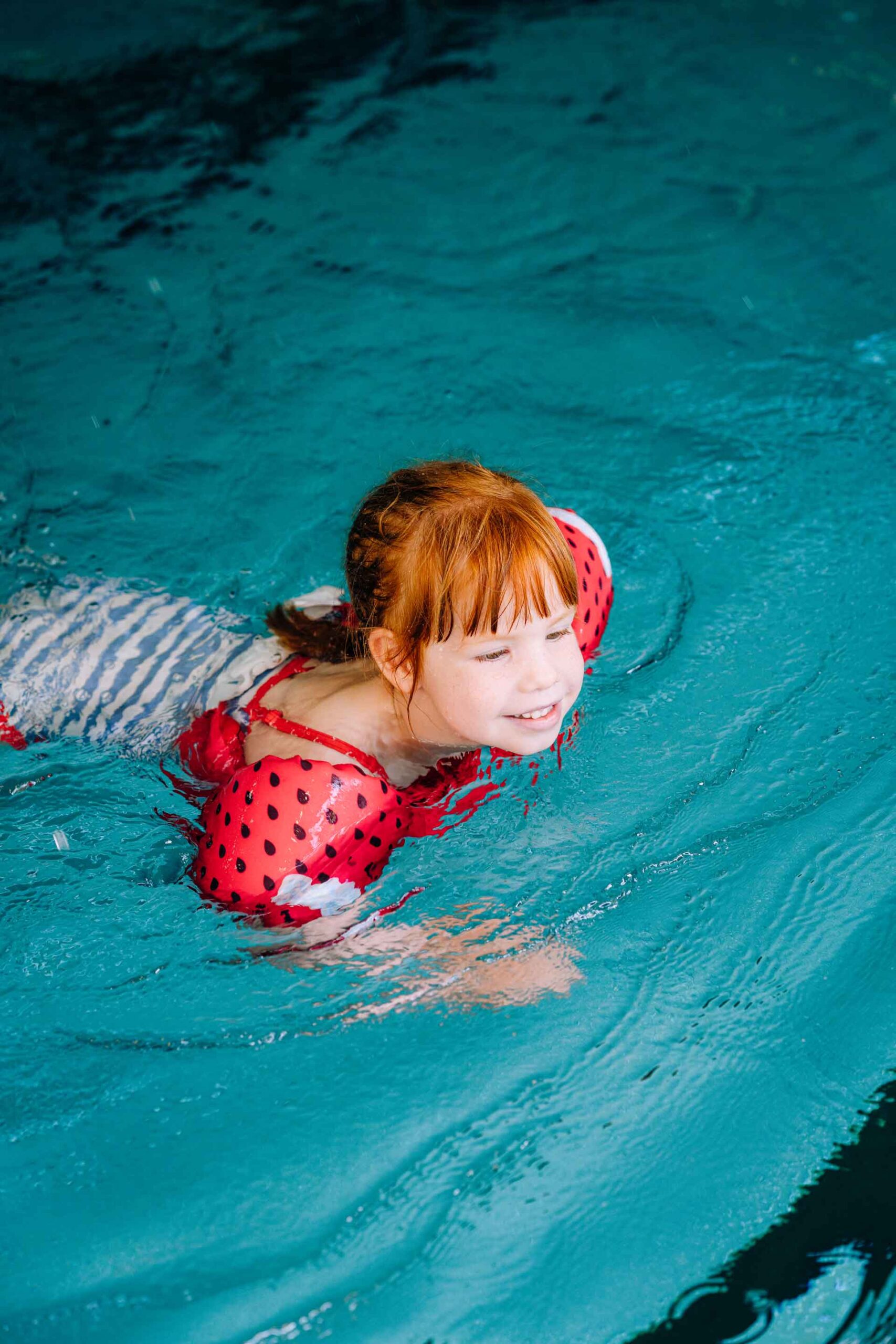 Rothaariges Kind mit roten Schwimmflügeln schwimmt beim Schwimmkurs durch ein türkisfarbenes Hallenbadbecken.