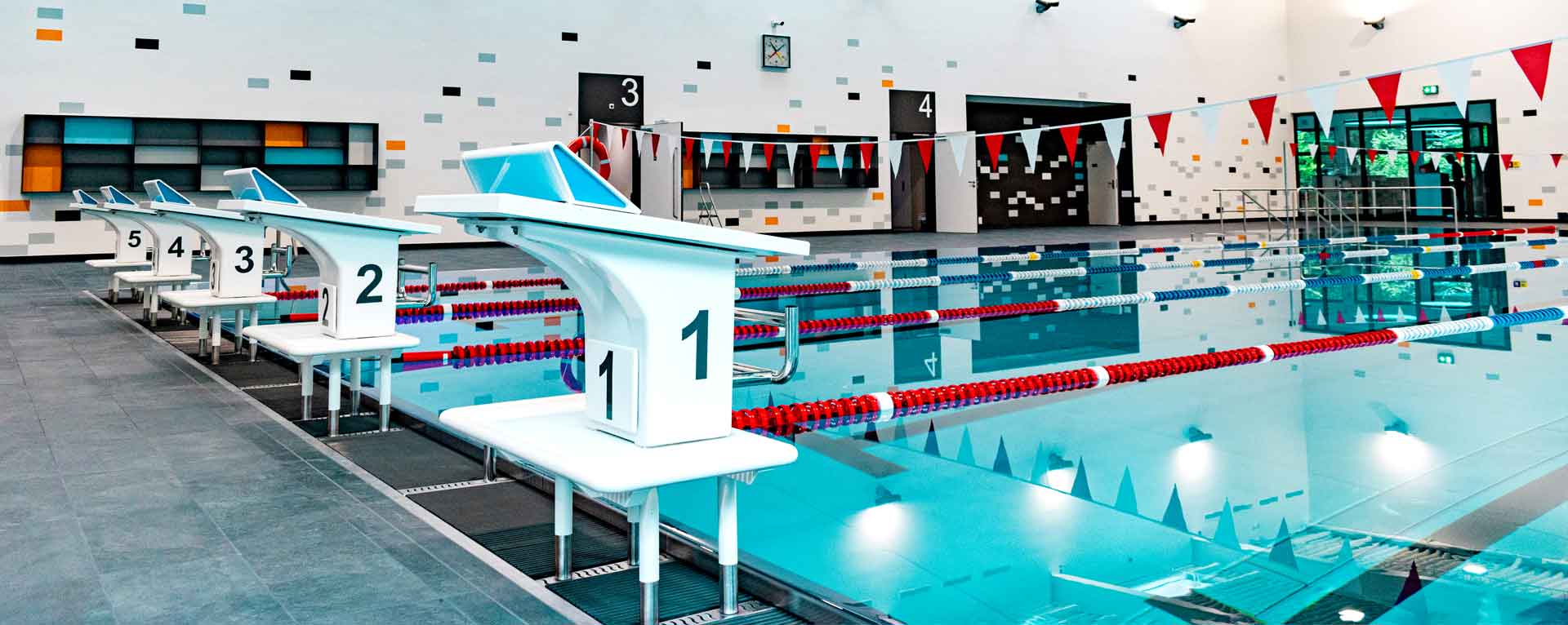 Weiße, nummerierte Startblöcke an einem leeren Schwimmbecken mit roten und blauen Bahnenleinen im Hallenbad.
